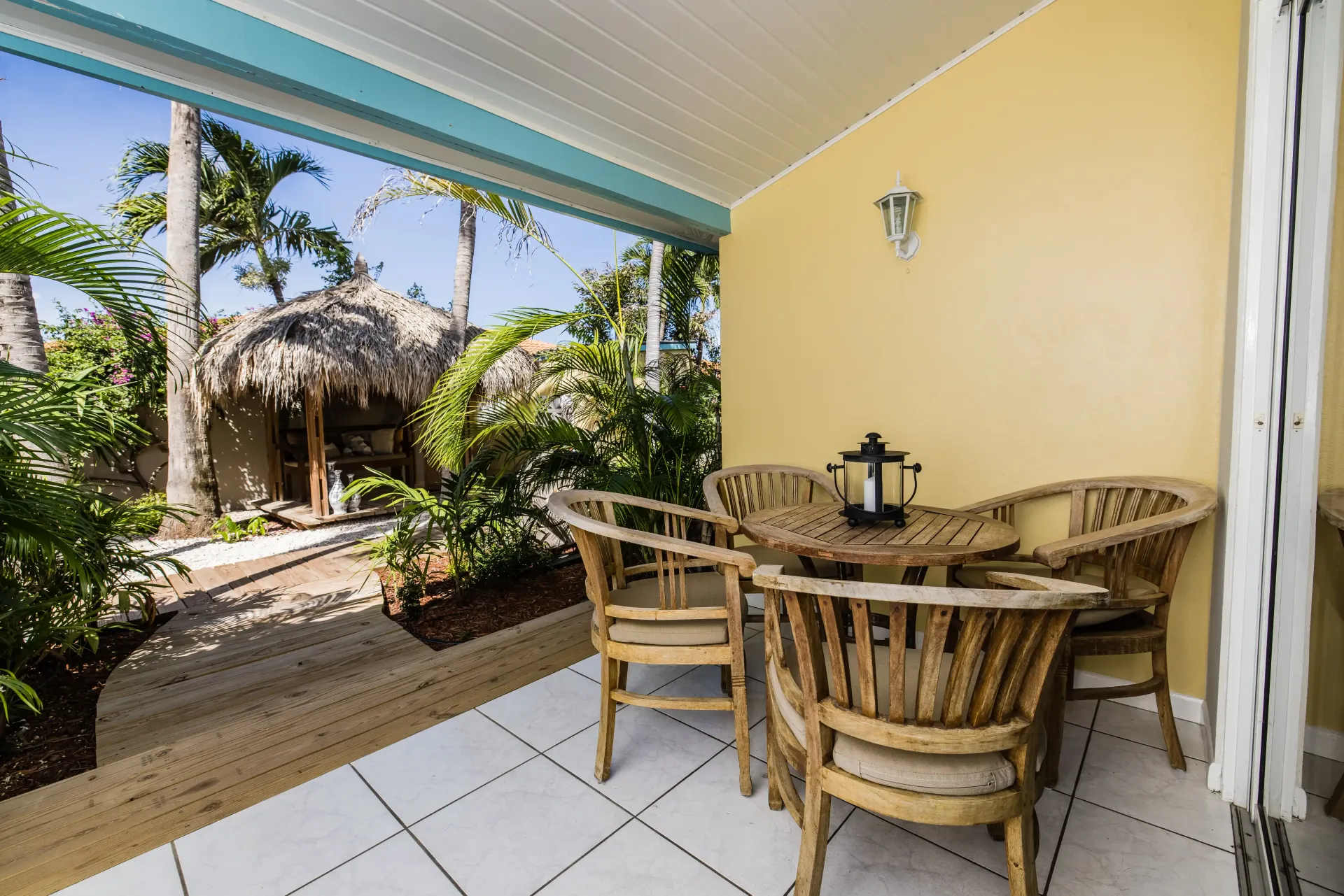 Patio with wicker furniture, view of thatched-roof building and tropical foliage. Yellow wall and blue ceiling.