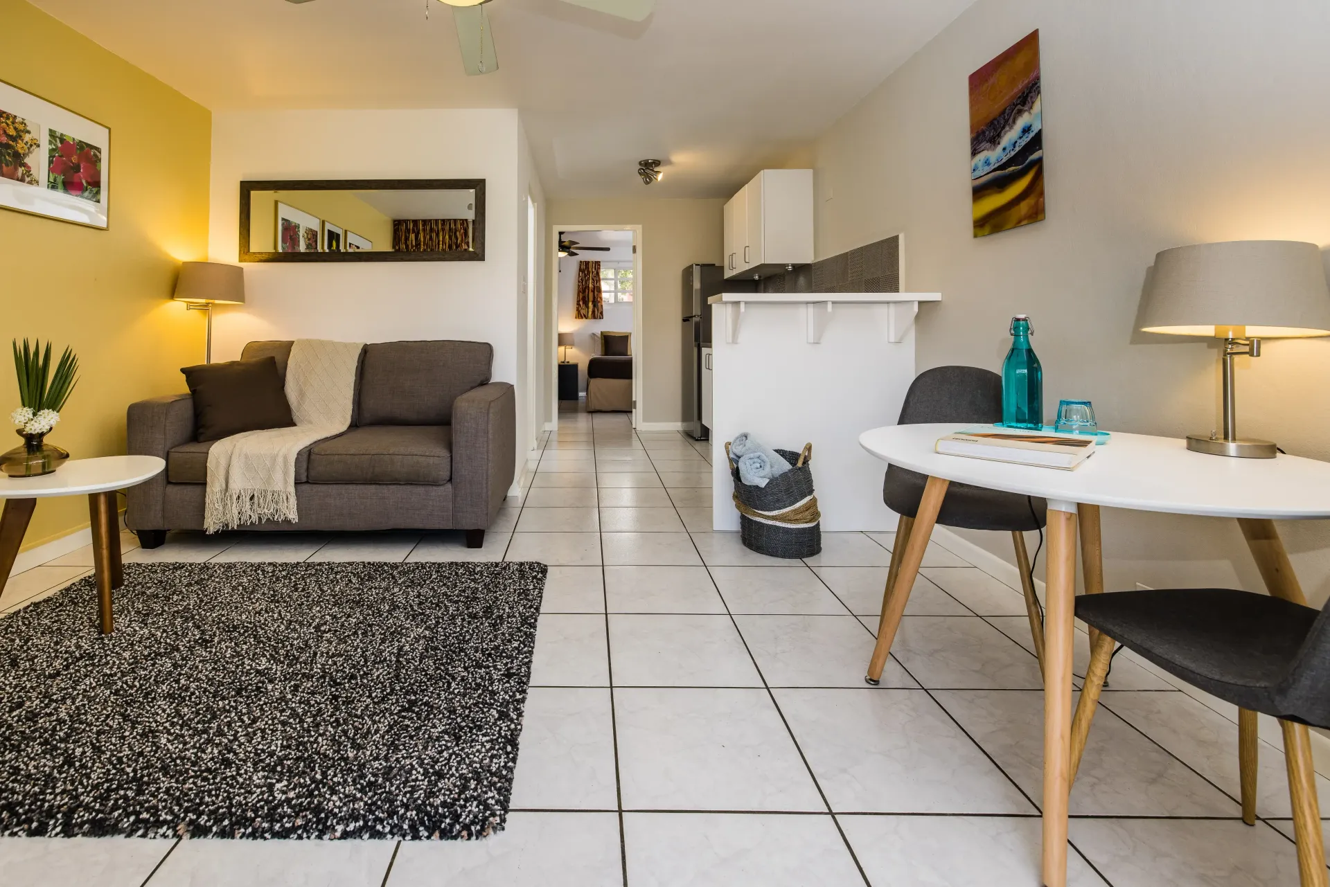 Living room with sofa, rug, table, chairs, and kitchen in the background. White and yellow walls.