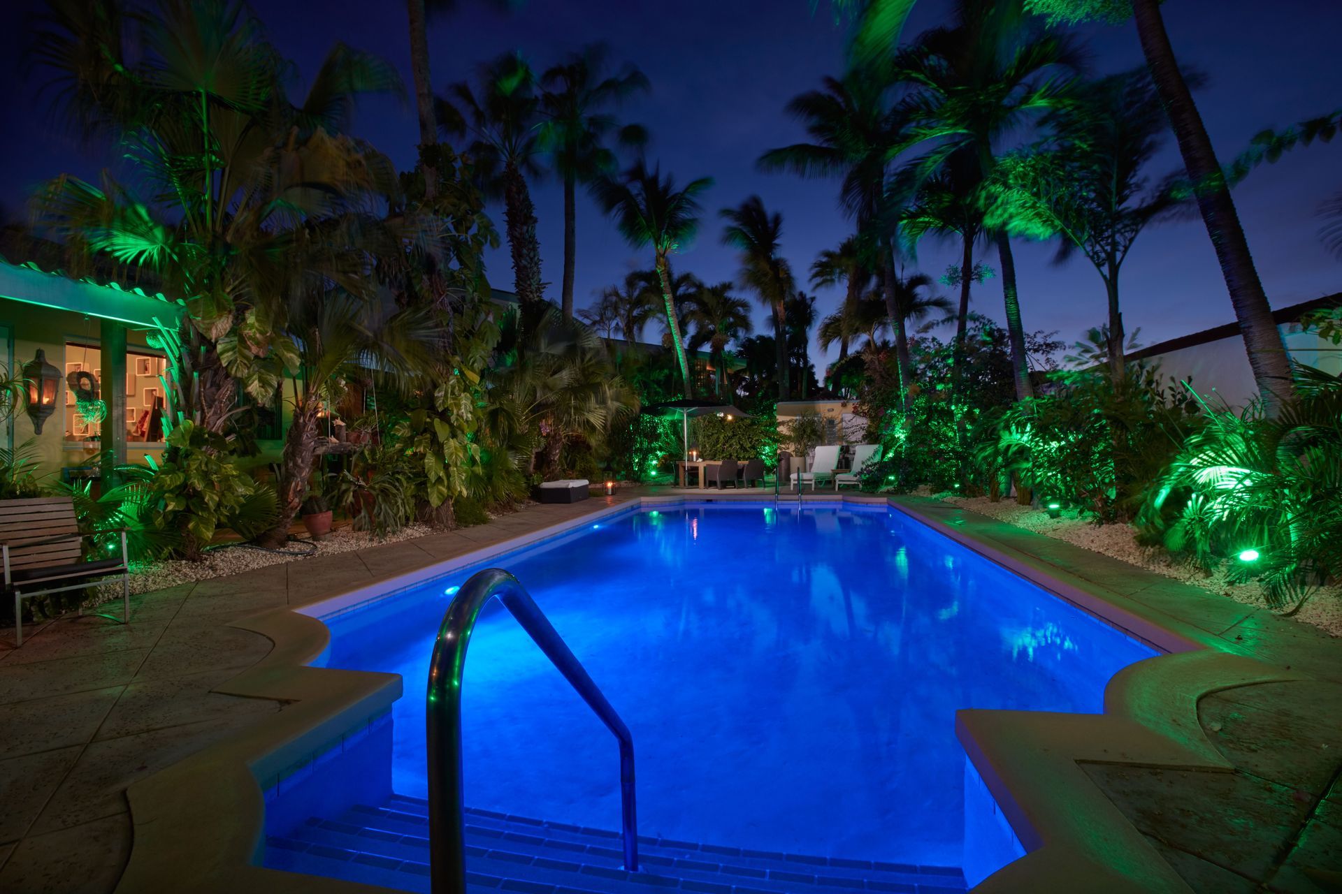 Nighttime pool with blue water, surrounded by tropical plants and illuminated by green lights.