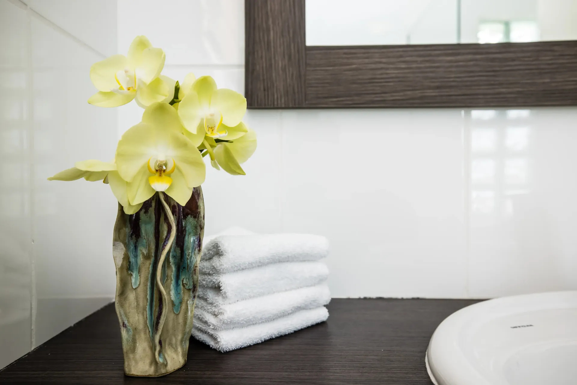 Yellow orchids in a textured vase, stacked white towels, and a sink on a dark counter in a bathroom with white tile.