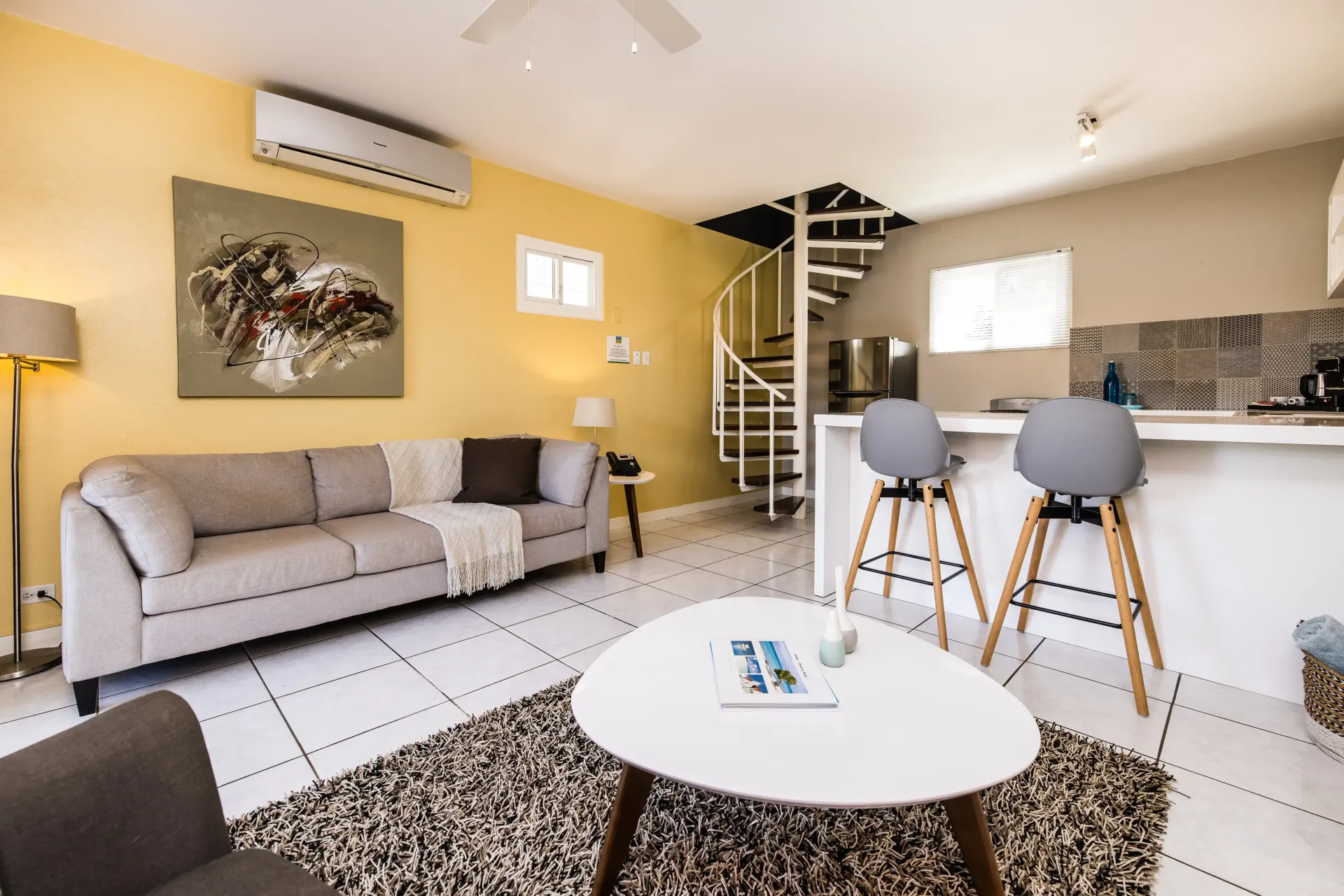 Living room with yellow walls, couch, spiral staircase, white counter with stools, and artwork.