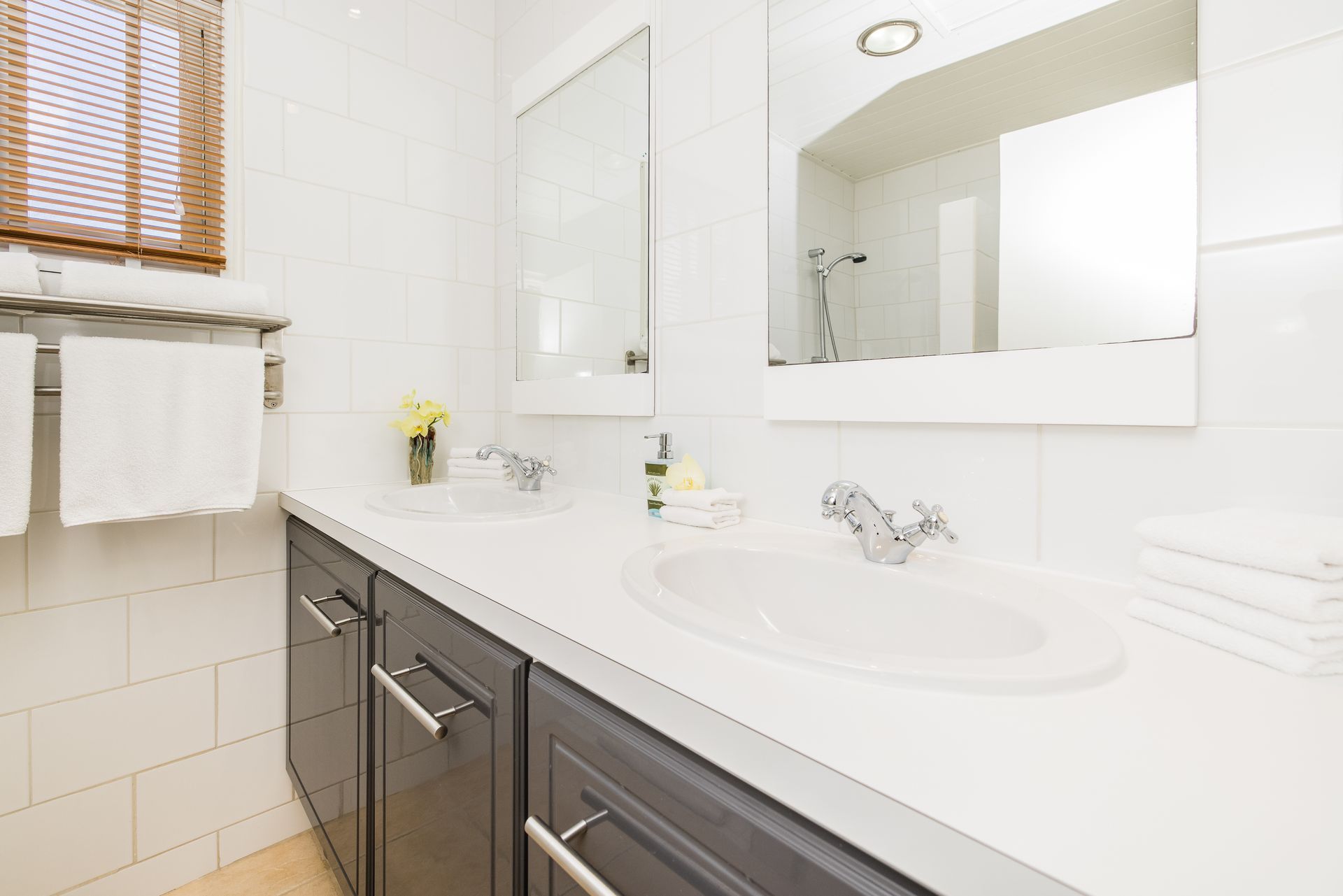 White bathroom with double sinks, gray cabinets, mirrors, and a window with blinds.