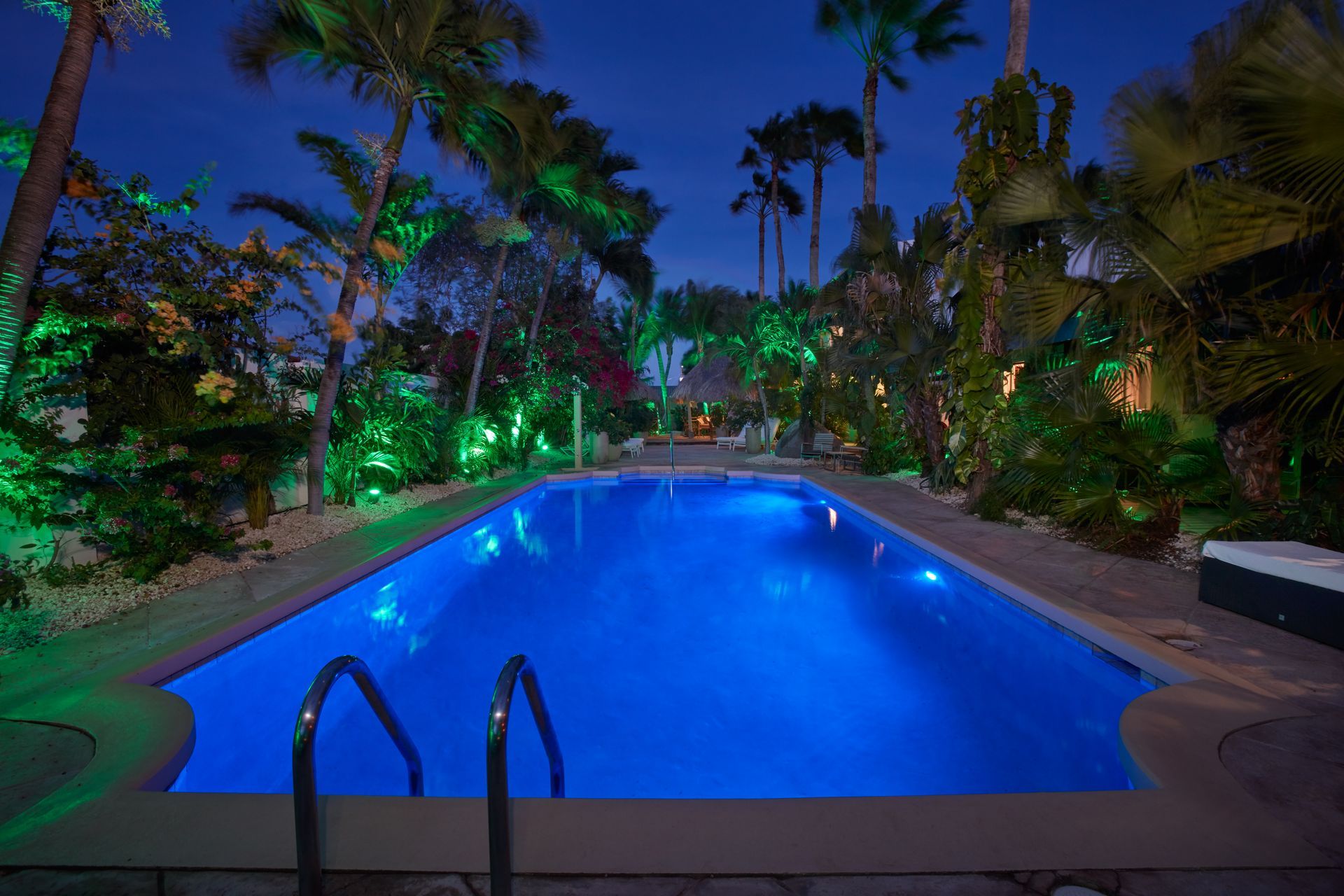 A nighttime pool scene with blue illuminated water, surrounded by lush greenery and palm trees.