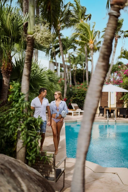 Couple walks near a pool, tropical setting. Woman in bikini, man in button-down shirt, both smiling. Palm trees, blue water.