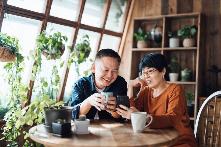 Two people smiling at a table, looking at a phone in a cozy café with plants and wooden shelves.
