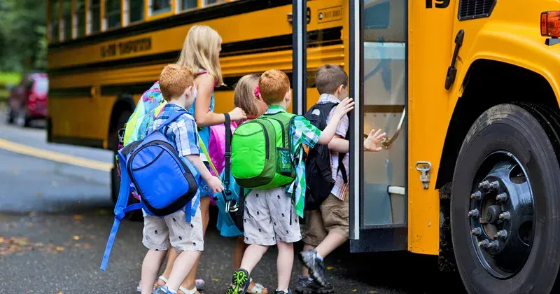 Children boarding a yellow school bus. Backpacks and a woman assist.