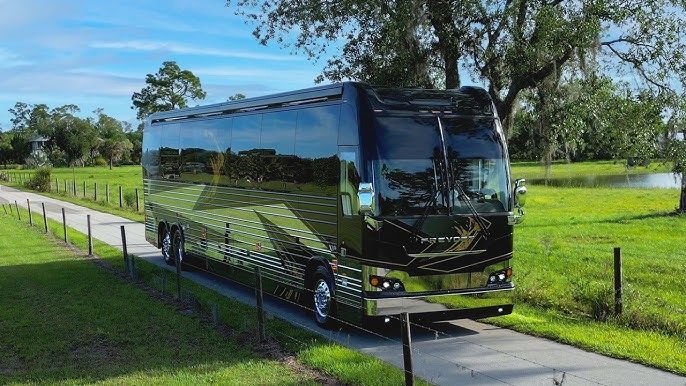 Black luxury RV on a paved road, green fields, trees in background.
