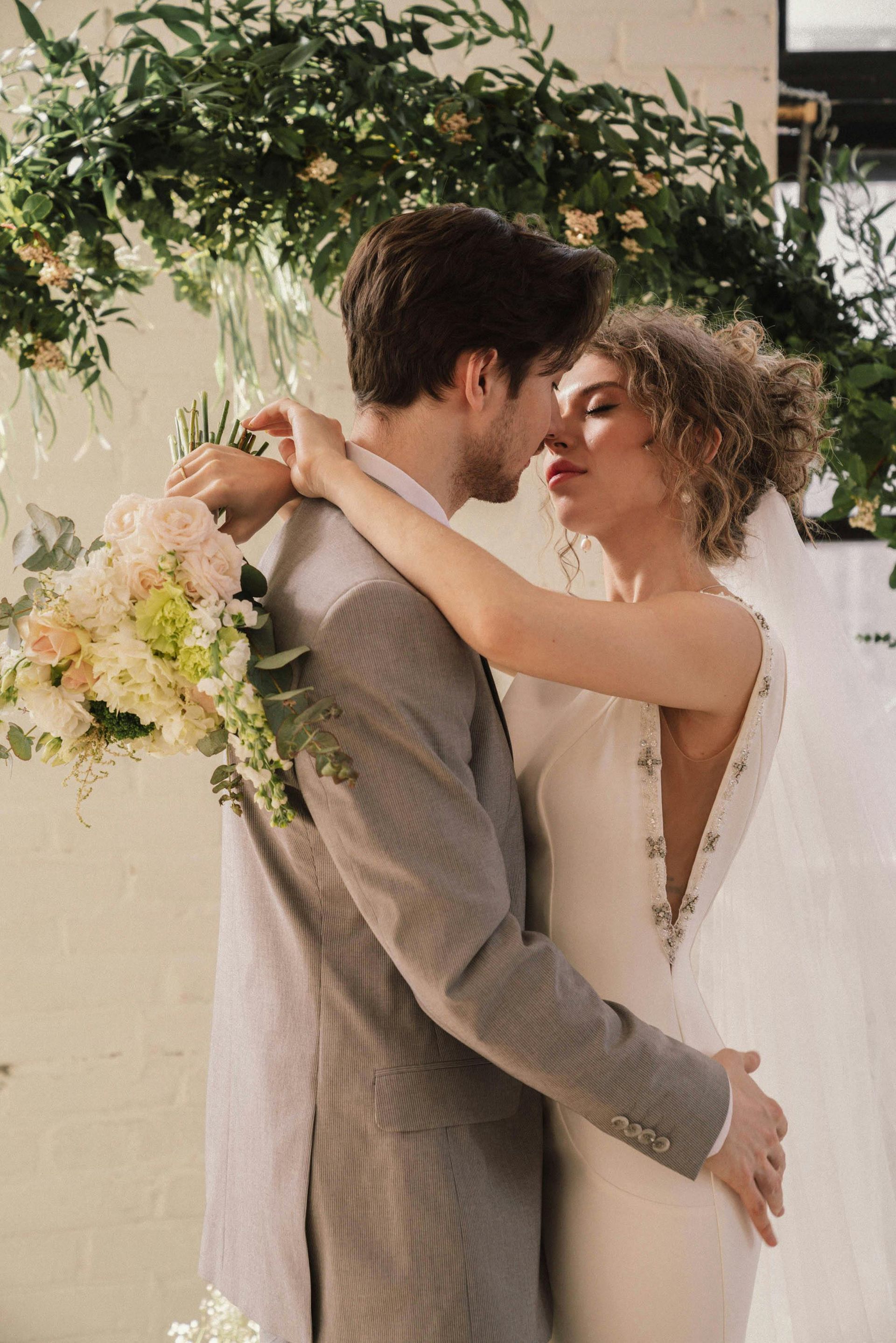 Bride and groom embrace, about to kiss. Wedding ceremony setting with floral arch, bouquet.