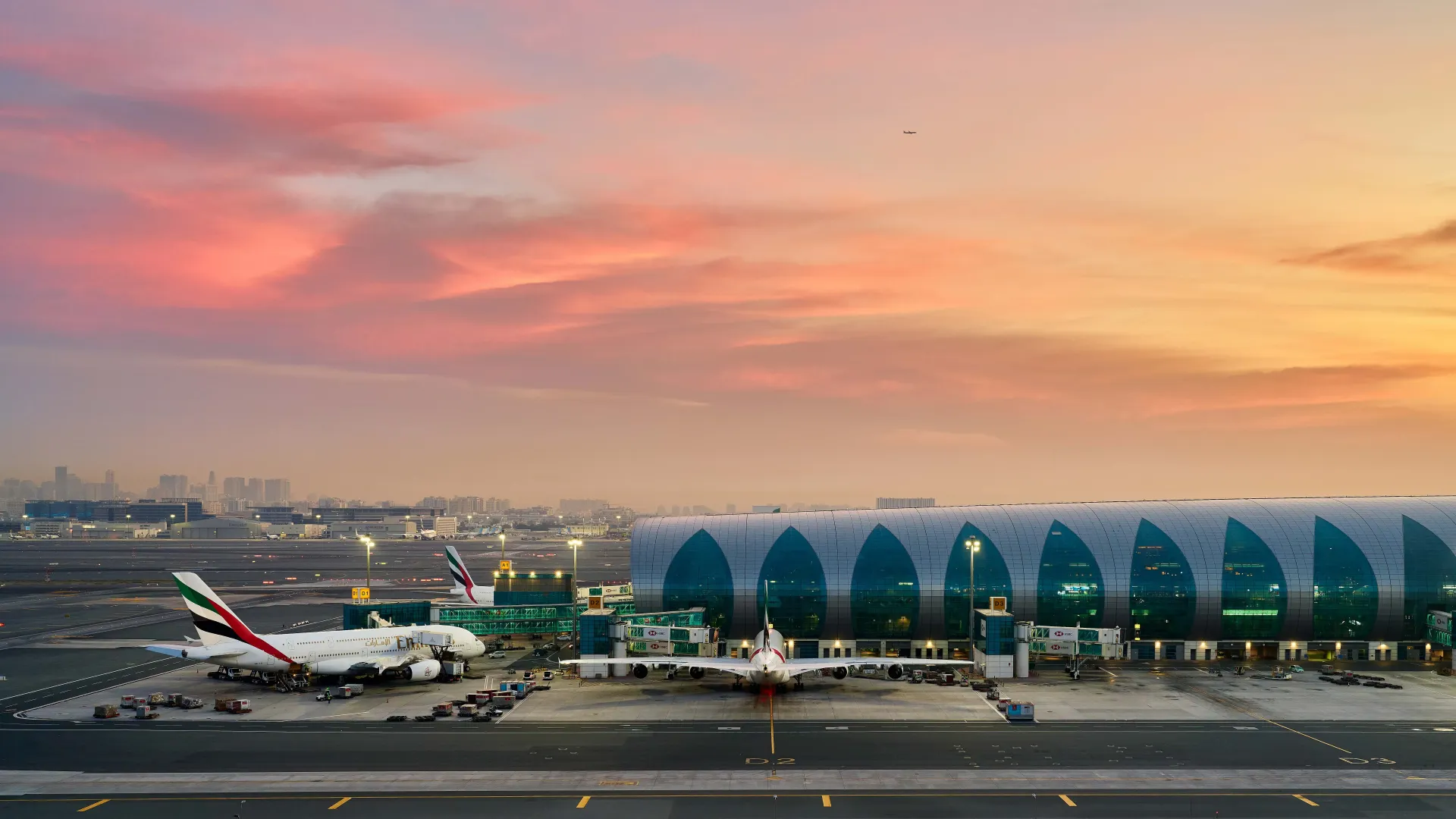 Airport terminal at sunset with airplanes on the tarmac; orange and pink sky.