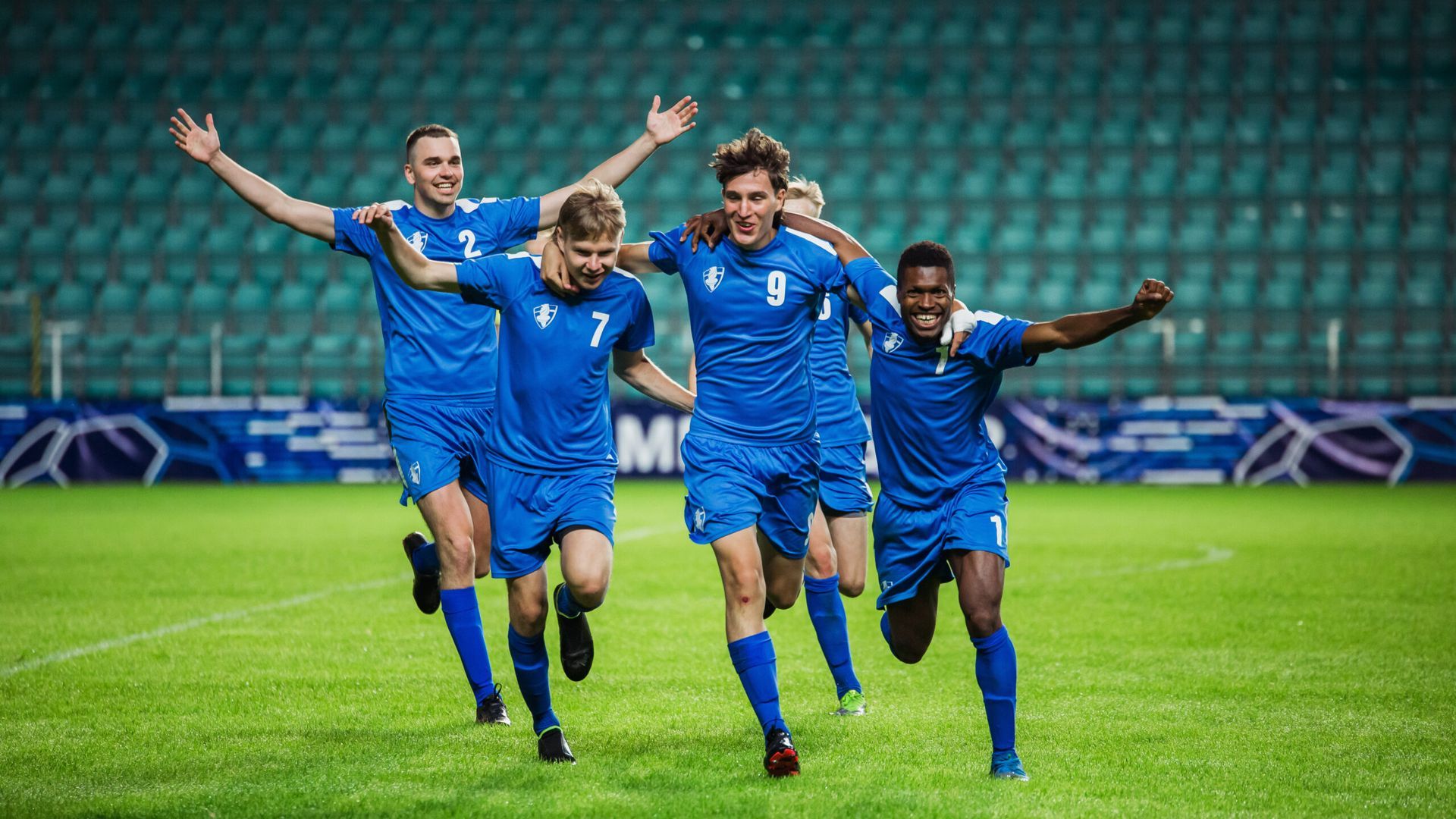 Soccer players in blue uniforms celebrating on a green field with arms raised.