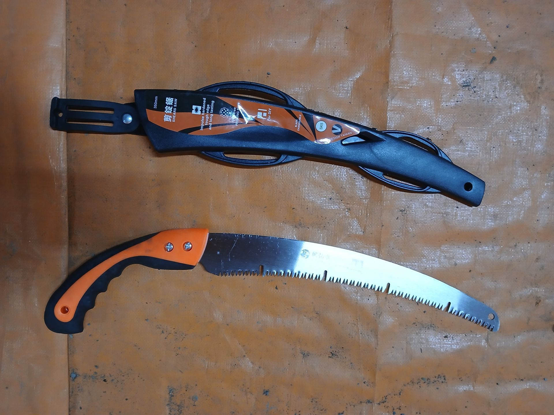 A folding hand saw with an orange and black handle, and a black sheath. The saw is on a textured orange surface. A folding hand saw with an orange and black handle, and a black sheath. The saw is on a textured orange surface.