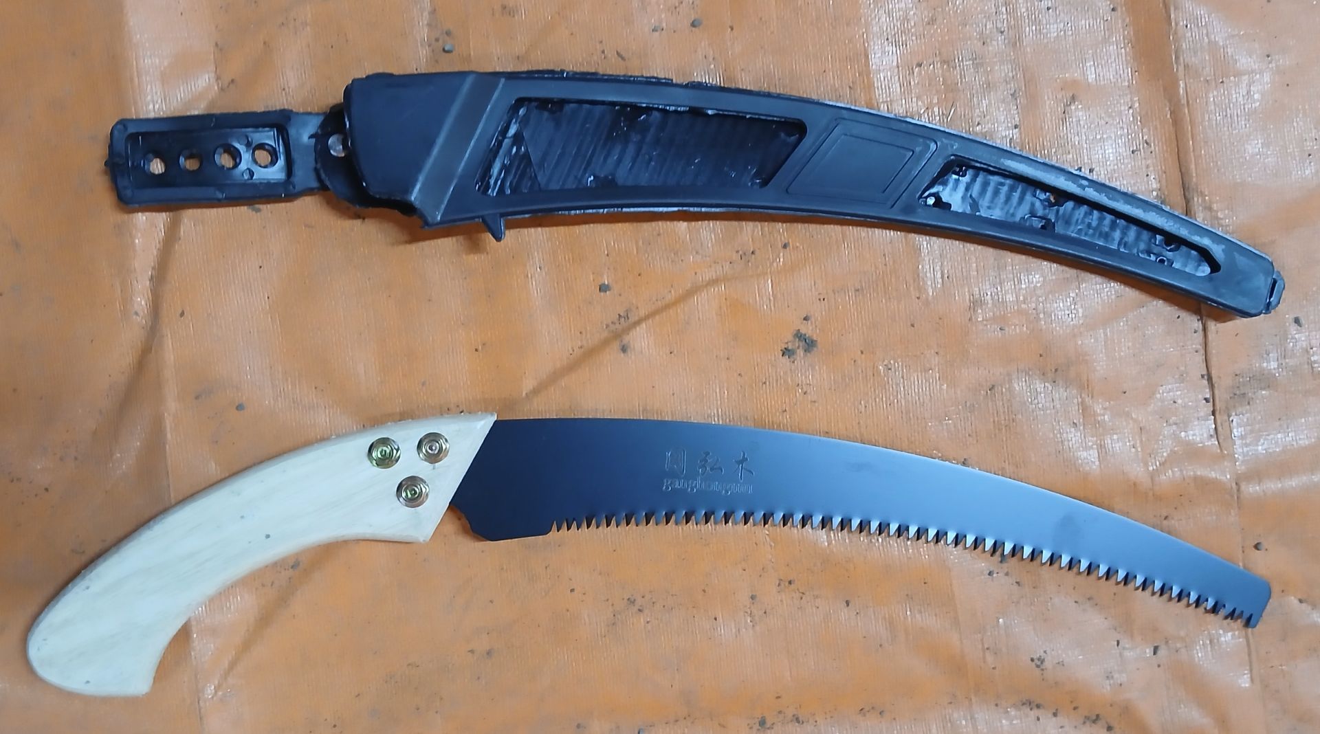 Black curved pruning saw with light wood handle, and matching black sheath on an orange surface. Black curved pruning saw with light wood handle, and matching black sheath on an orange surface.