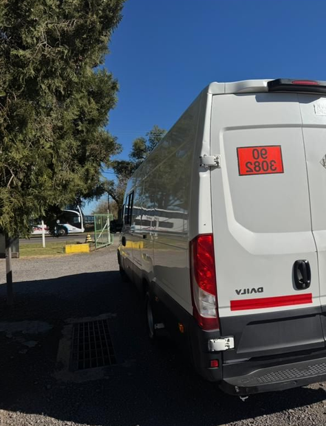 Furgoneta blanca aparcada al aire libre junto a un árbol, con un cartel rojo en la parte trasera y una zona vallada delante.