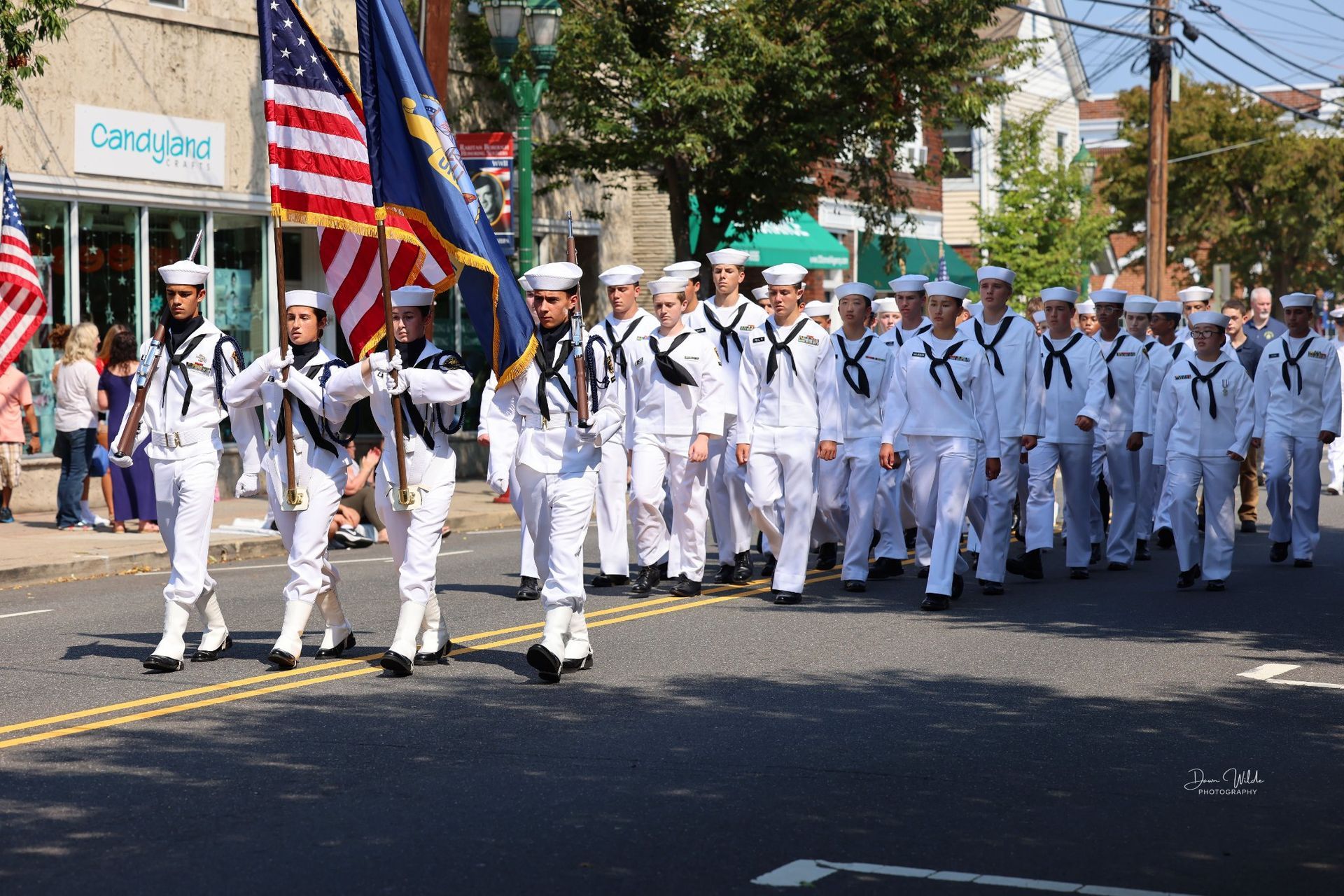 A group of men in white uniforms are marching down a street holding flags.