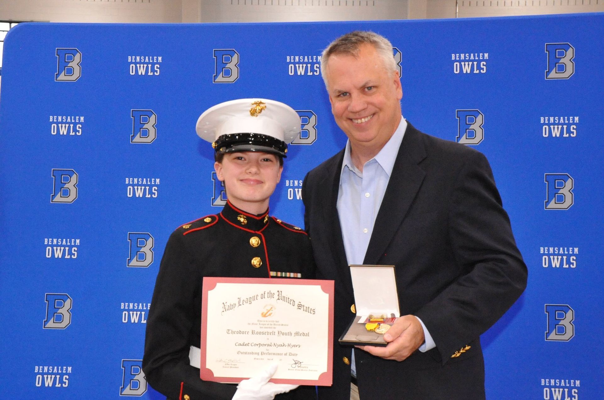 A man in a suit stands next to a girl in a marine uniform holding a certificate