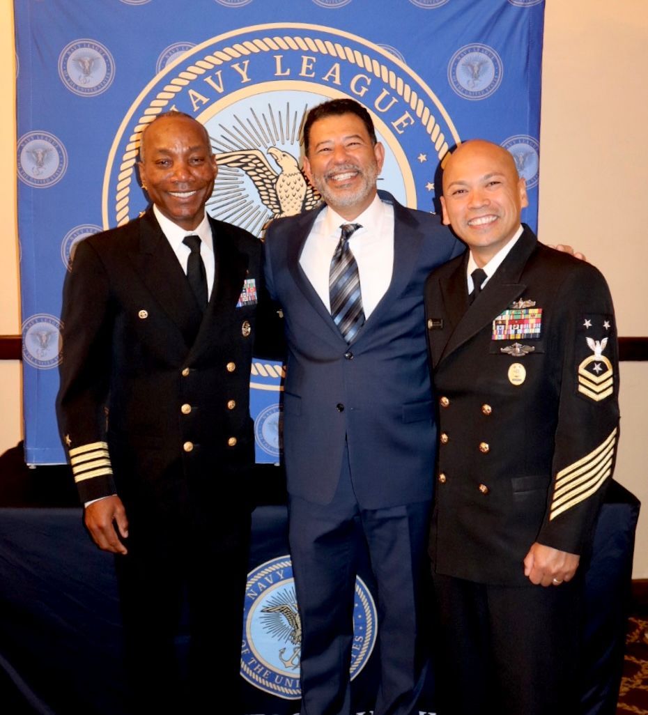 Three men are posing for a picture in front of a navy league banner