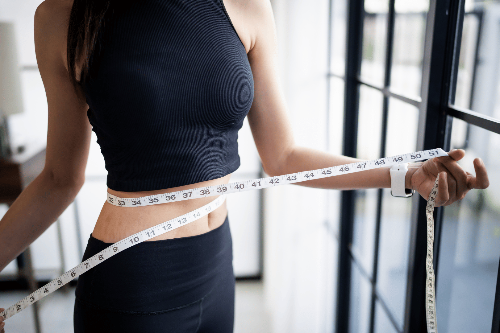 Woman in black athletic wear measuring waist with a tape measure.