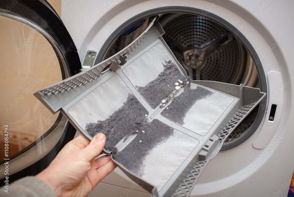 Person holding lint filter from dryer, full of grey lint, in front of open dryer door.