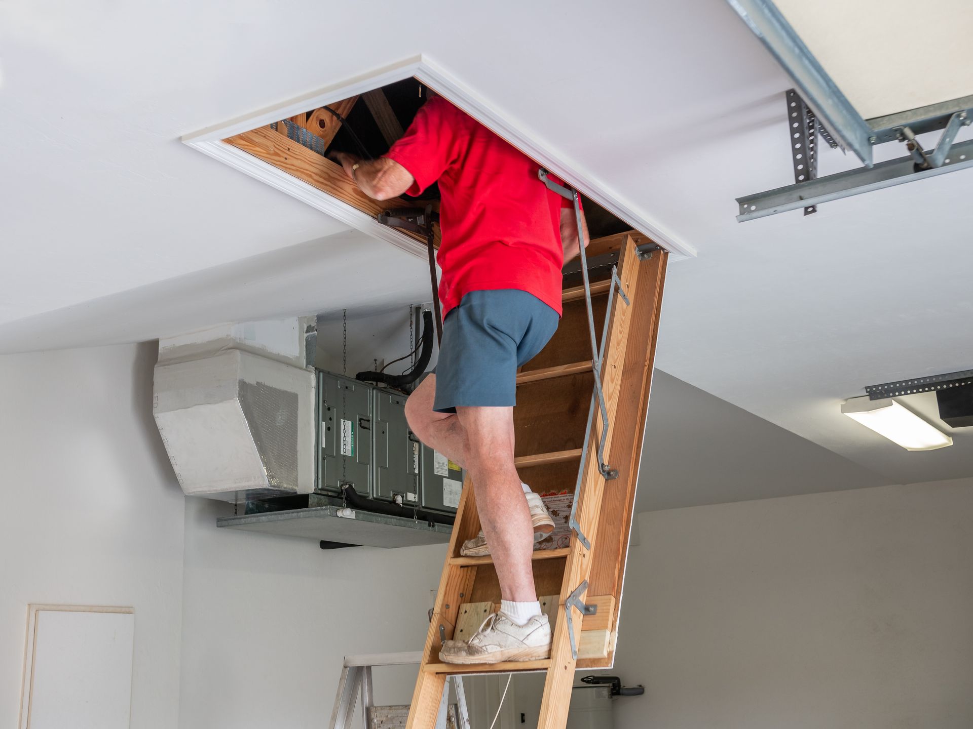Man in red shirt and shorts climbing into attic via pull-down ladder in a garage.