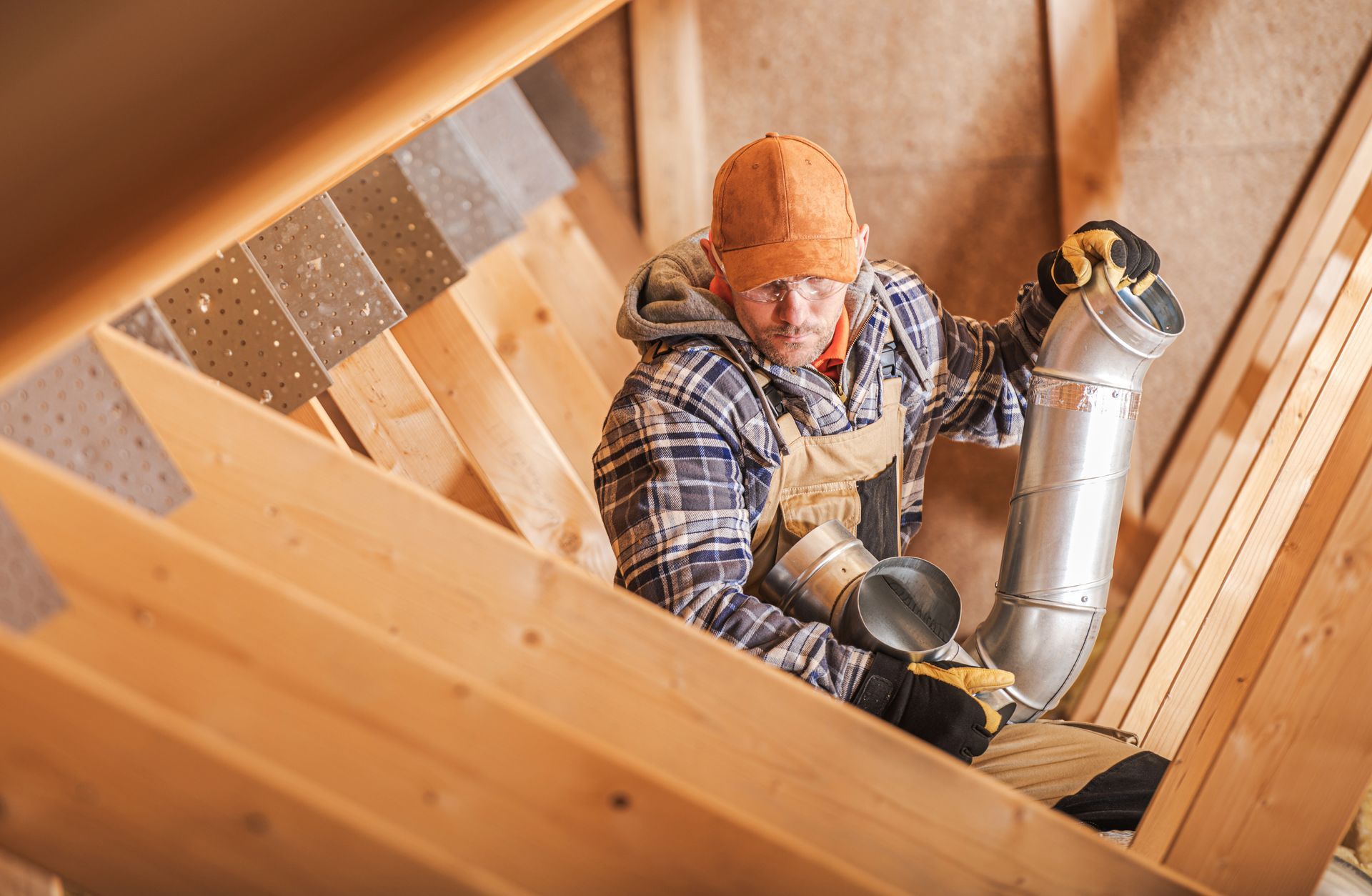 Man in an attic installing ductwork; wearing a hat, gloves, and overalls.