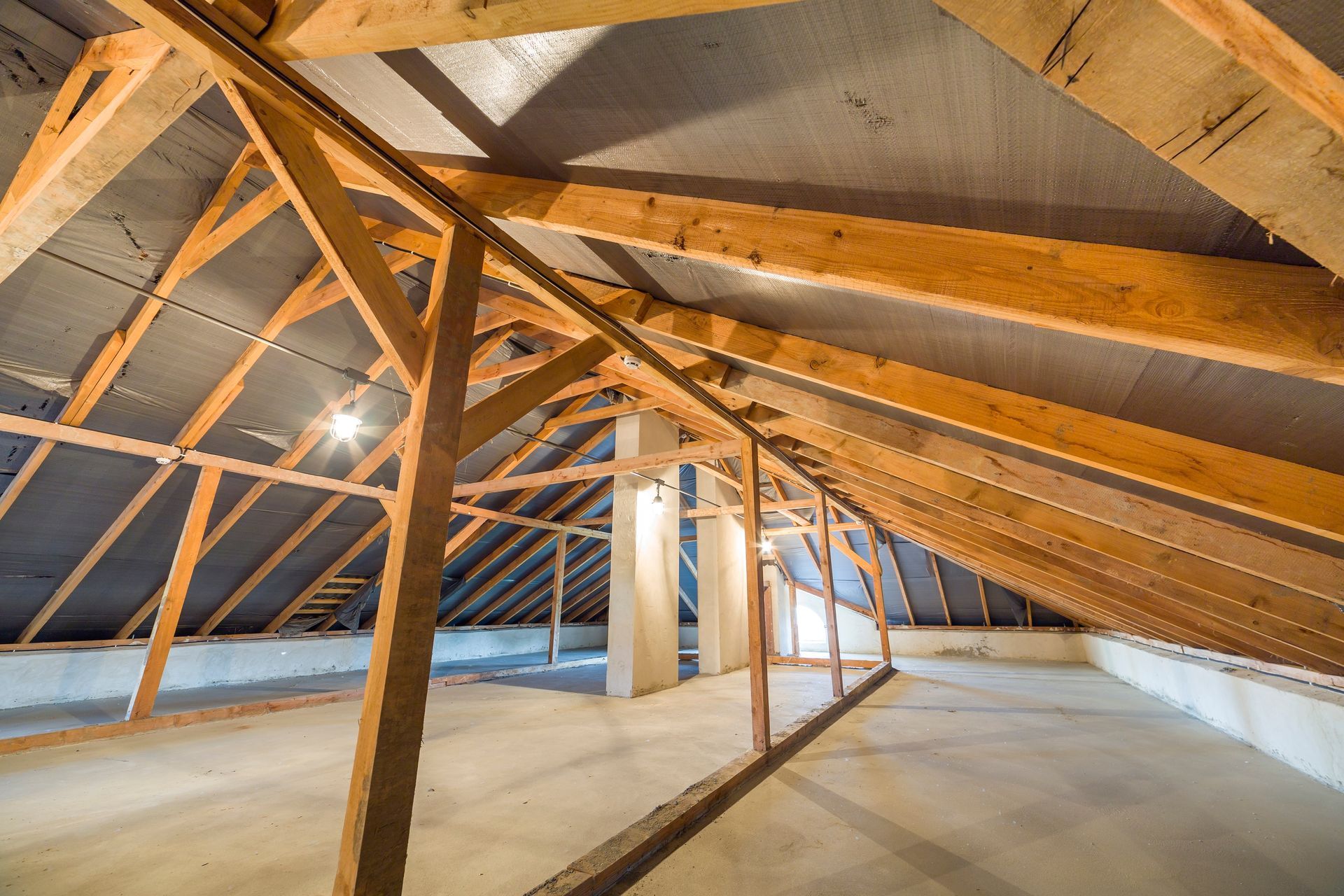 Wooden attic structure with rafters and beams. Gray insulation is attached, and the floor is concrete.