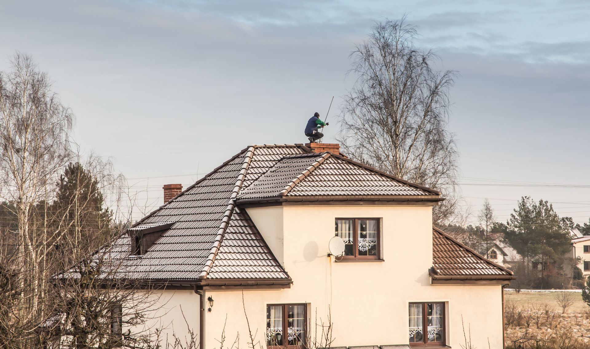 Person on a snowy rooftop of a two-story house, inspecting a chimney. Overcast sky.
