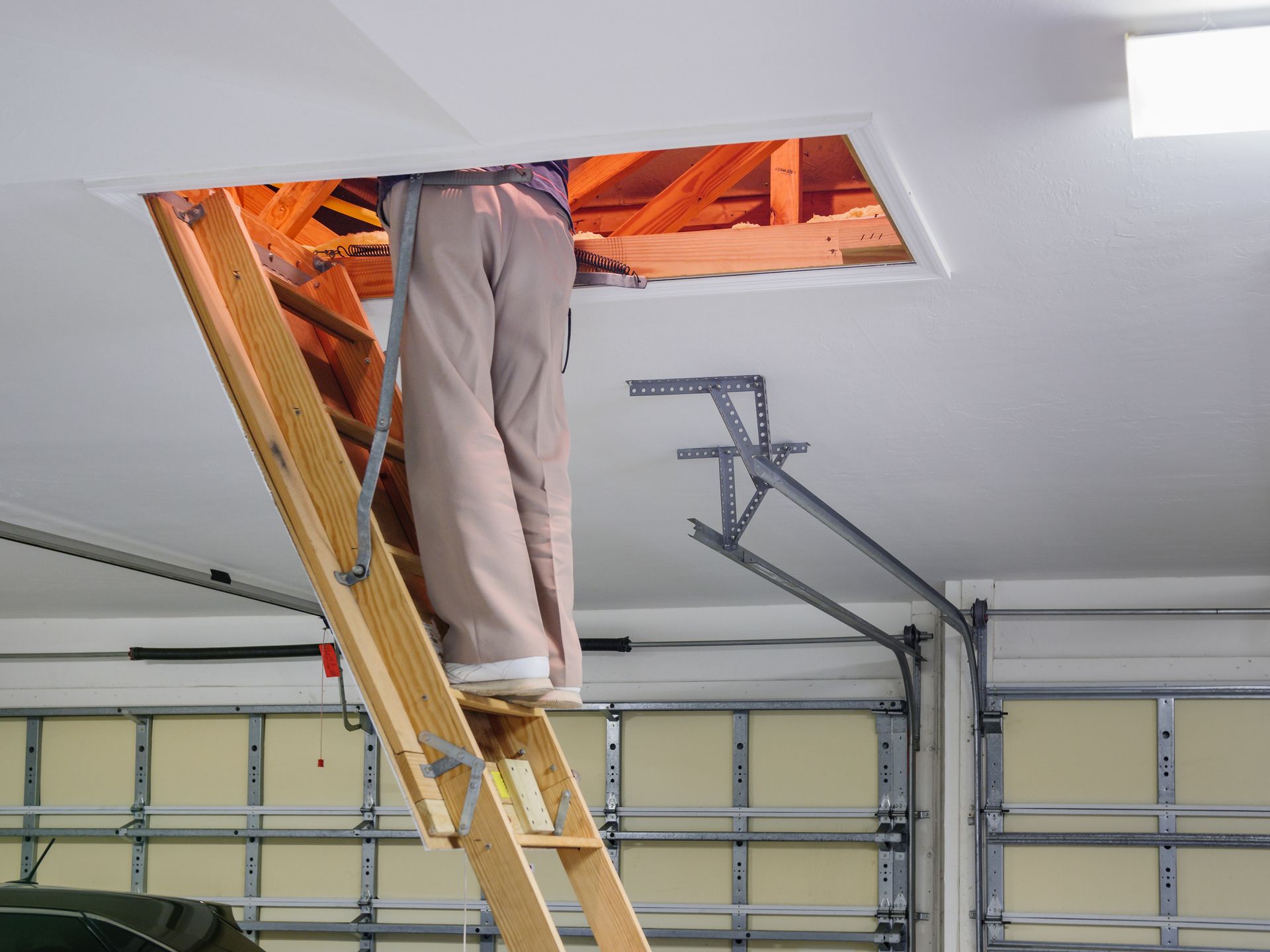 Person climbing a wooden attic ladder into an attic opening in a garage ceiling.