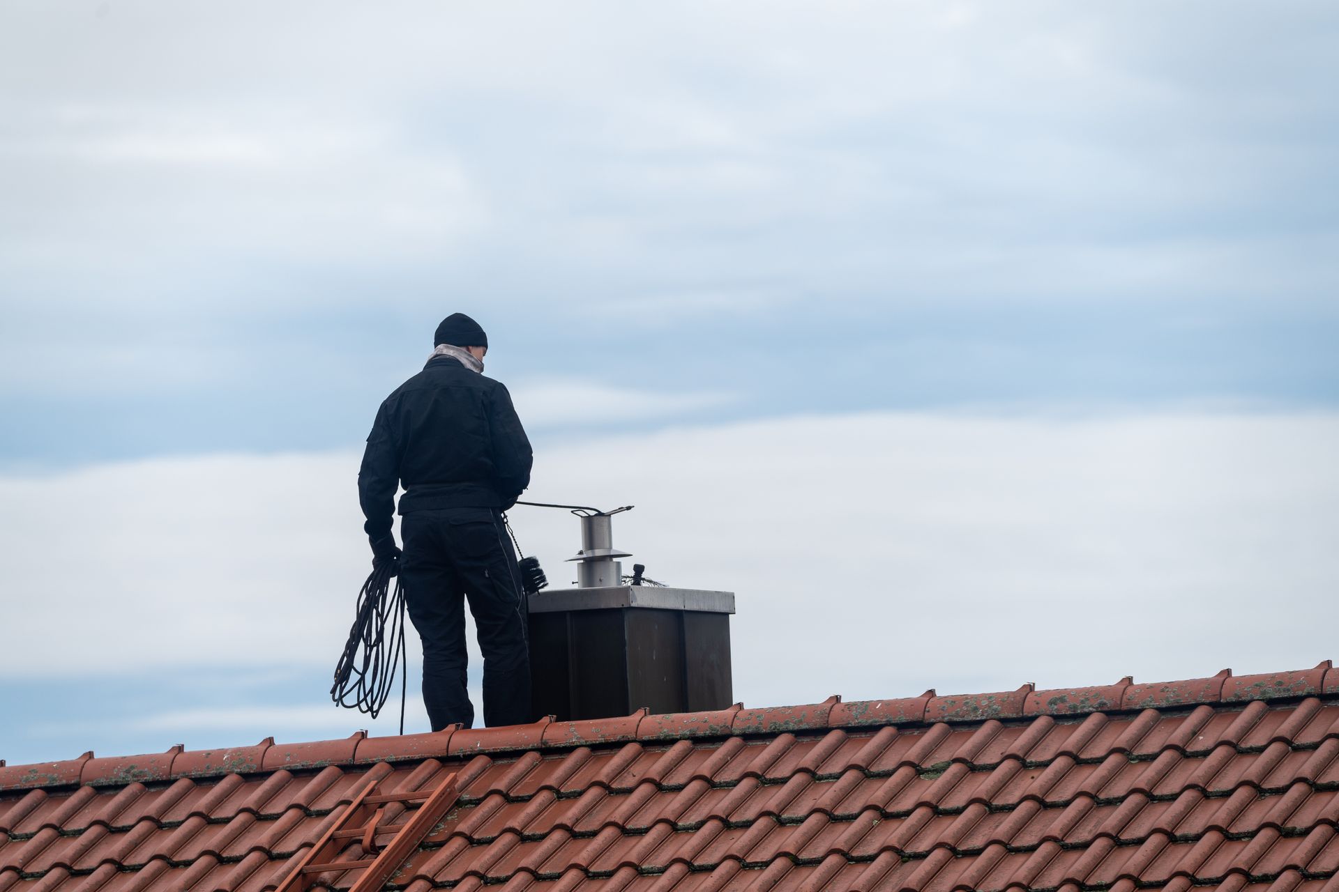 A person on a red tile roof, near a chimney, against a cloudy sky. They are wearing black and holding tools.
