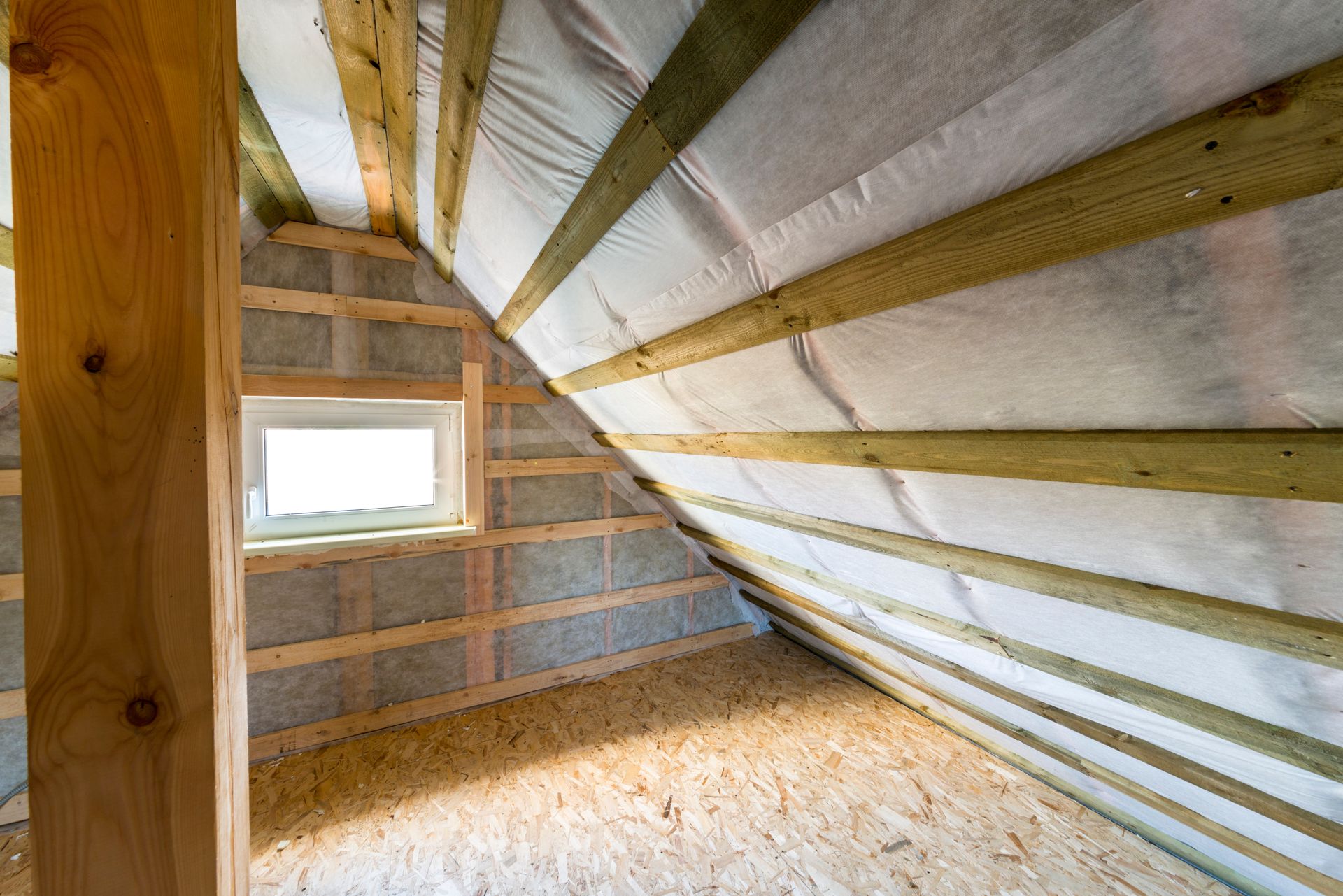 Interior of a roof attic under construction, showing wooden beams, insulation, and a small window.