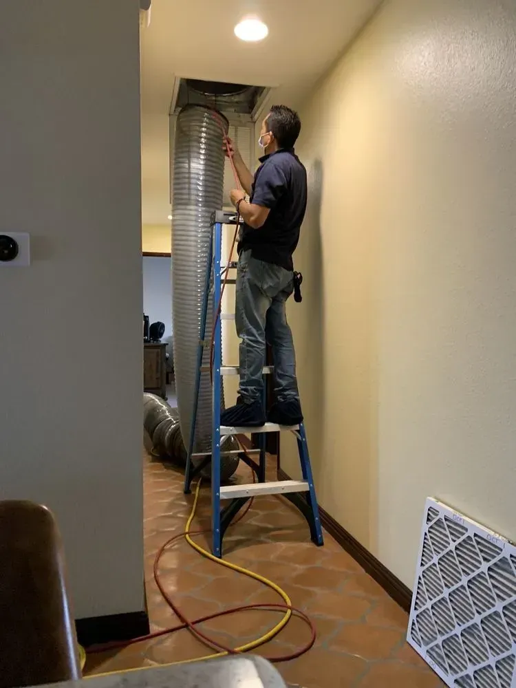 Person on ladder inspecting ductwork in a hallway; a new filter lies on the floor.