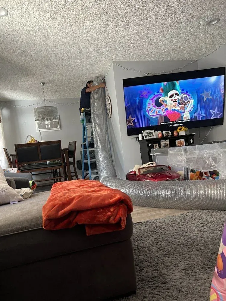 Person on ladder installing flexible ductwork indoors, near TV and couch.