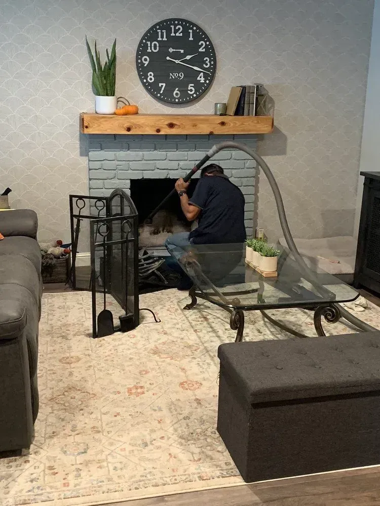 Man cleaning a fireplace with a vacuum. Fireplace is gray brick, with a wooden mantel and clock above.