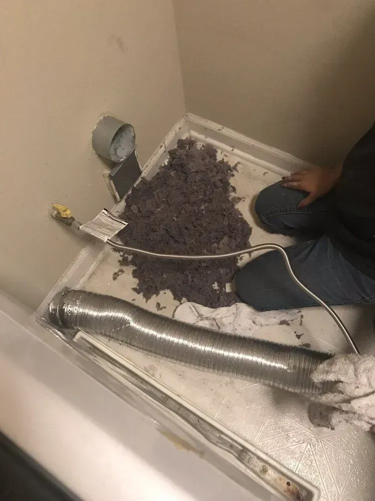Laundry room with person kneeling. Large pile of lint on floor near dryer vent and flexible ductwork.