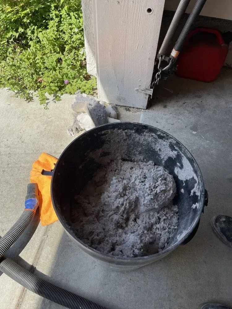 Black bucket filled with gray debris next to a white pillar on a concrete surface.
