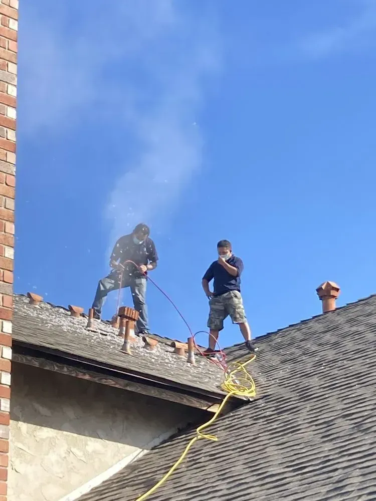 Two people on a roof working on a chimney. One sprays water, the other stands beside him. Bright blue sky.