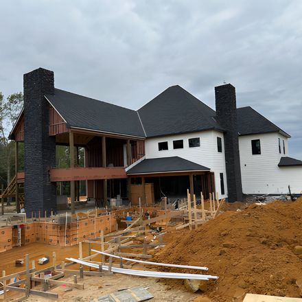 House under construction, with black shingled roof, white exterior, and stone chimneys, with dirt and lumber in front.