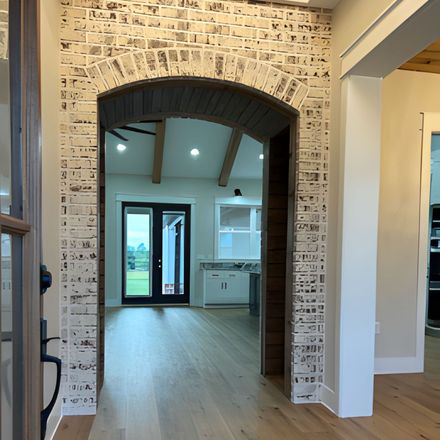 Arched doorway with whitewashed brick, leading to a room with wood beams, light wood floors, and black doors.