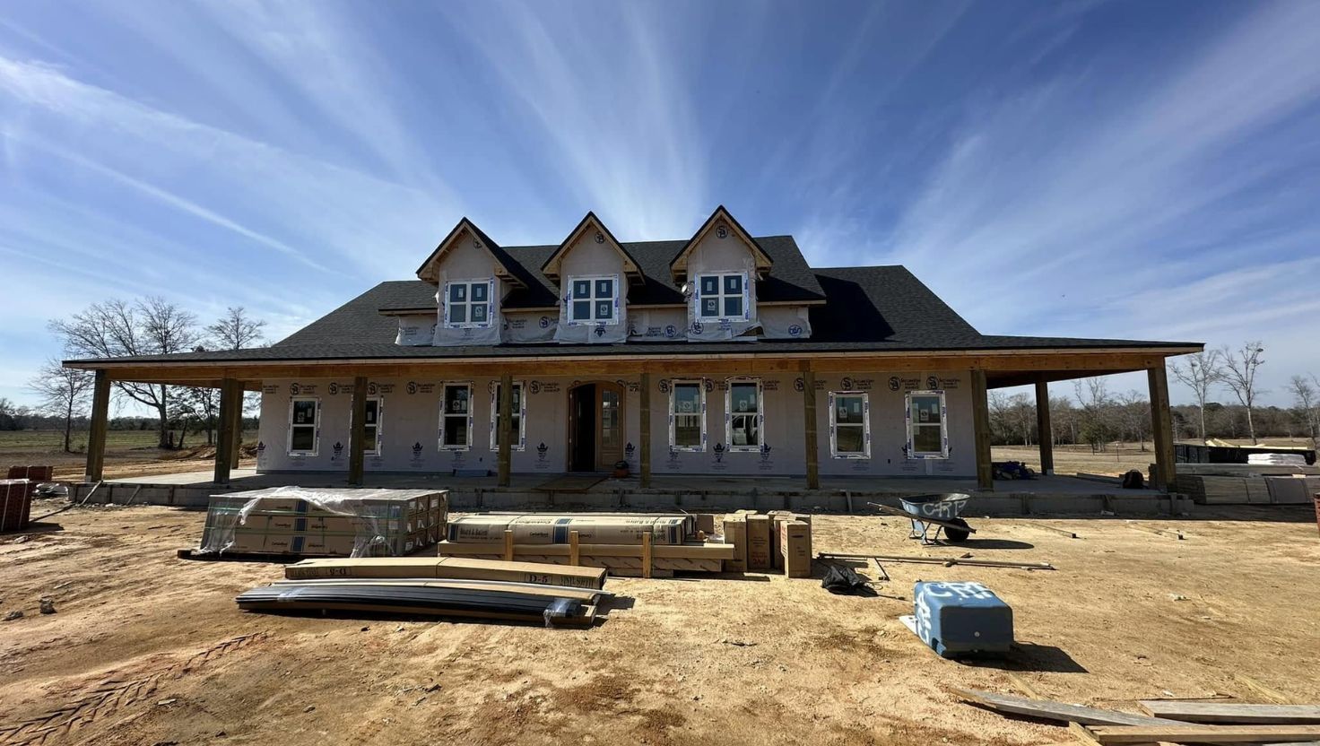 House under construction with porch, gabled roof, and stacked building materials. Sunny outdoor setting.