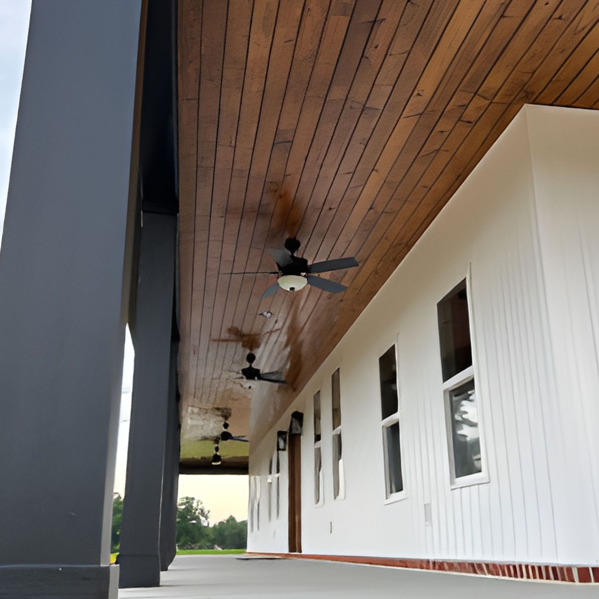 Exterior of a building with white walls, dark gray columns, and a wooden ceiling with ceiling fans.