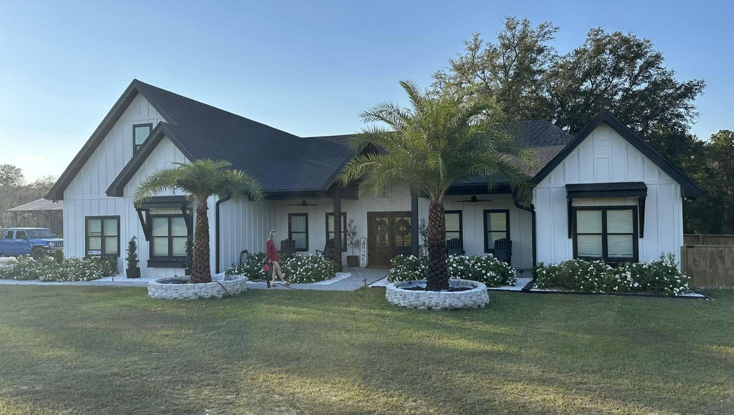 White farmhouse with black trim, palm trees, and green lawn under a blue sky.