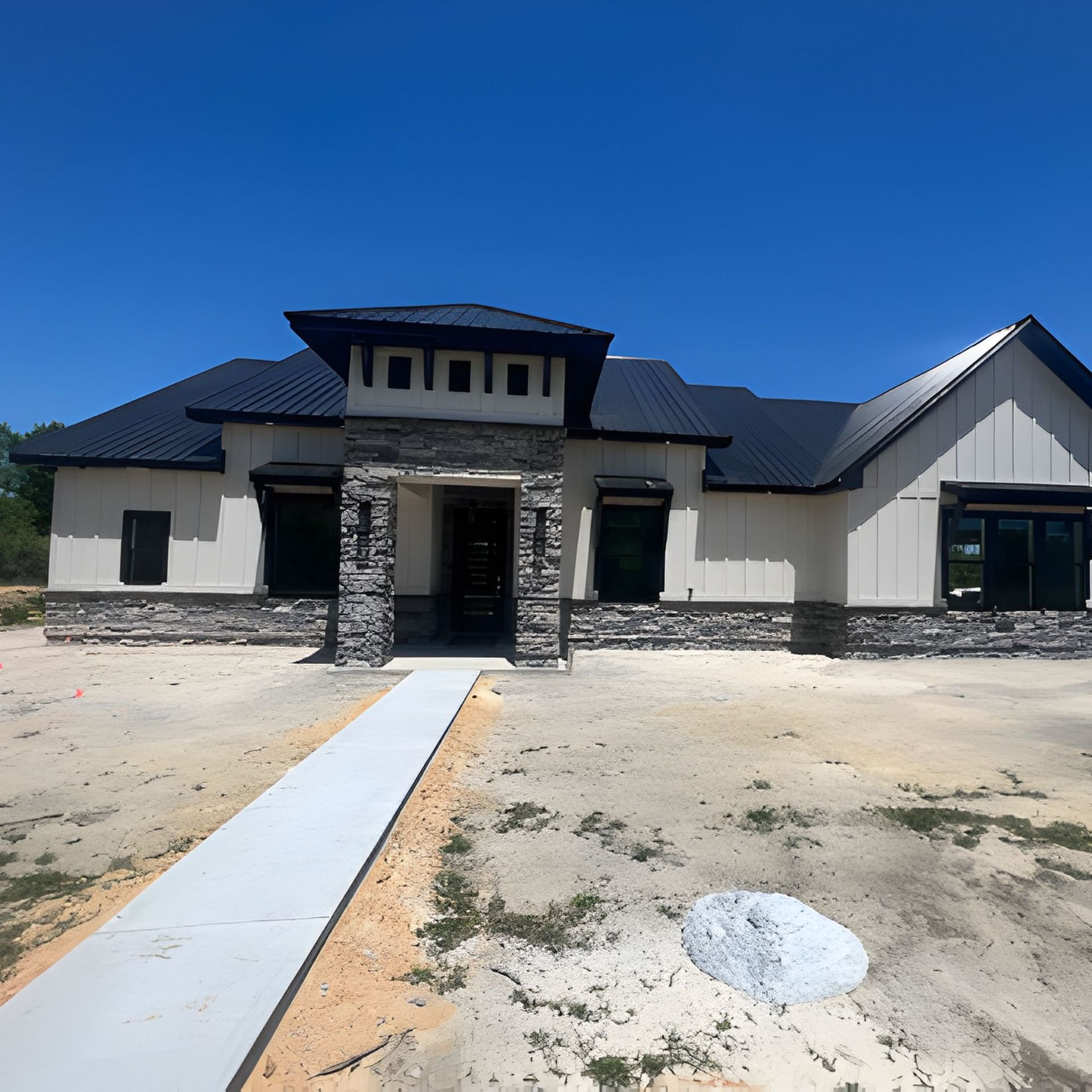 Modern house under construction with white siding, stone accents, black roof, and clear blue sky.