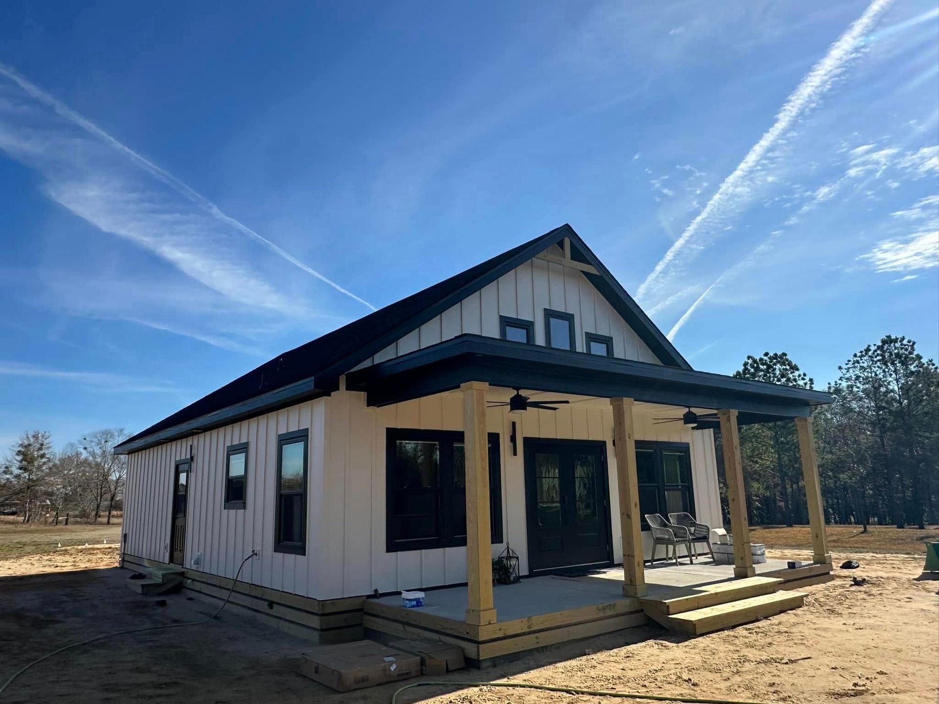 A white house with a black roof and a porch is sitting on top of a dirt field.