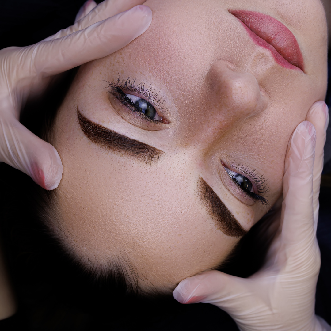 Woman's face close-up, brow procedure, hands with gloves holding her head. Dark eyebrows, full lips, light skin.