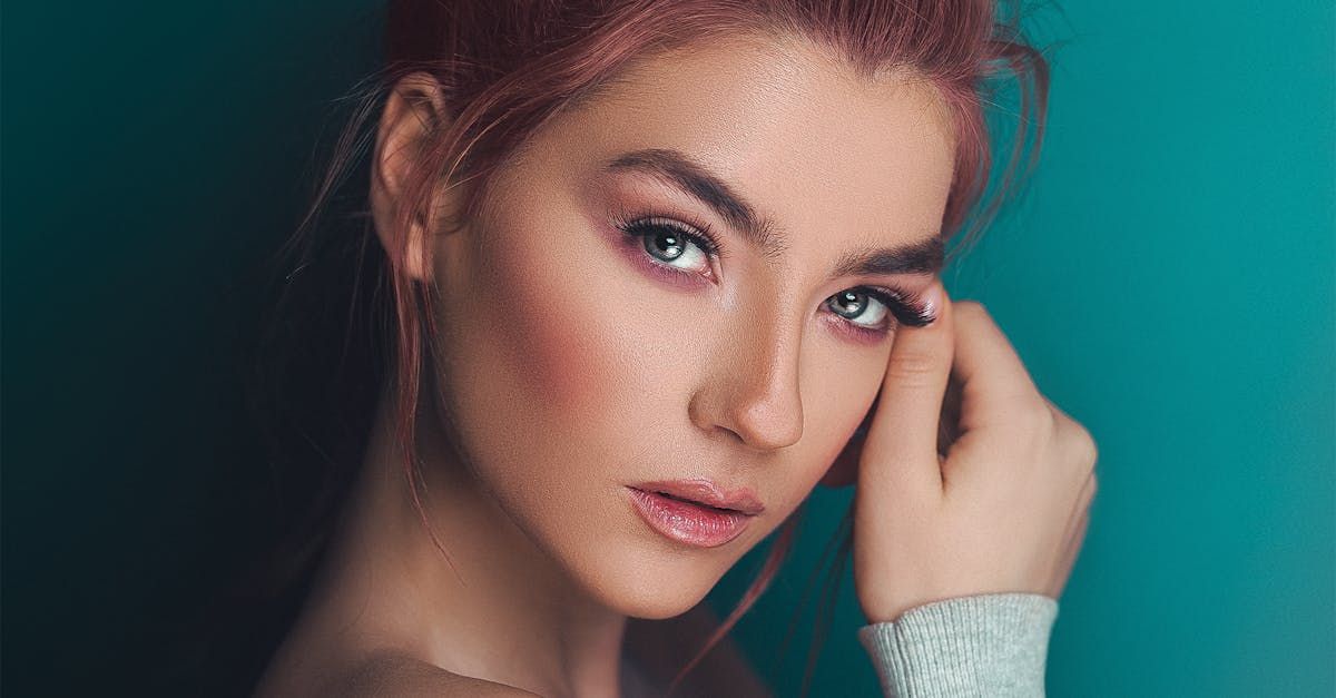 Woman with pink hair and makeup, hand on face, looking at camera against a teal background.