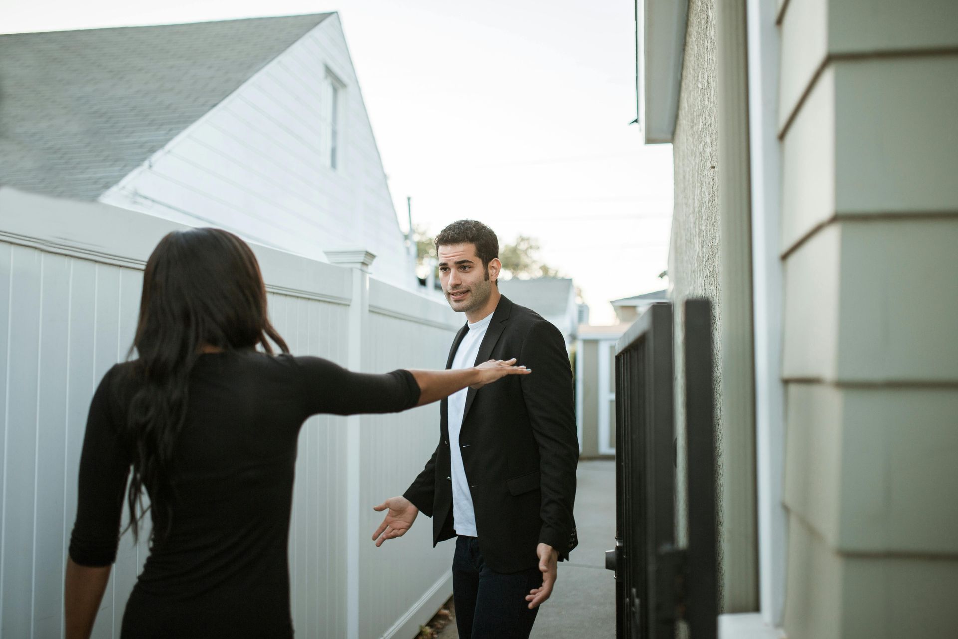 Woman gesturing, man with open arms, standing in narrow space between fences and buildings.