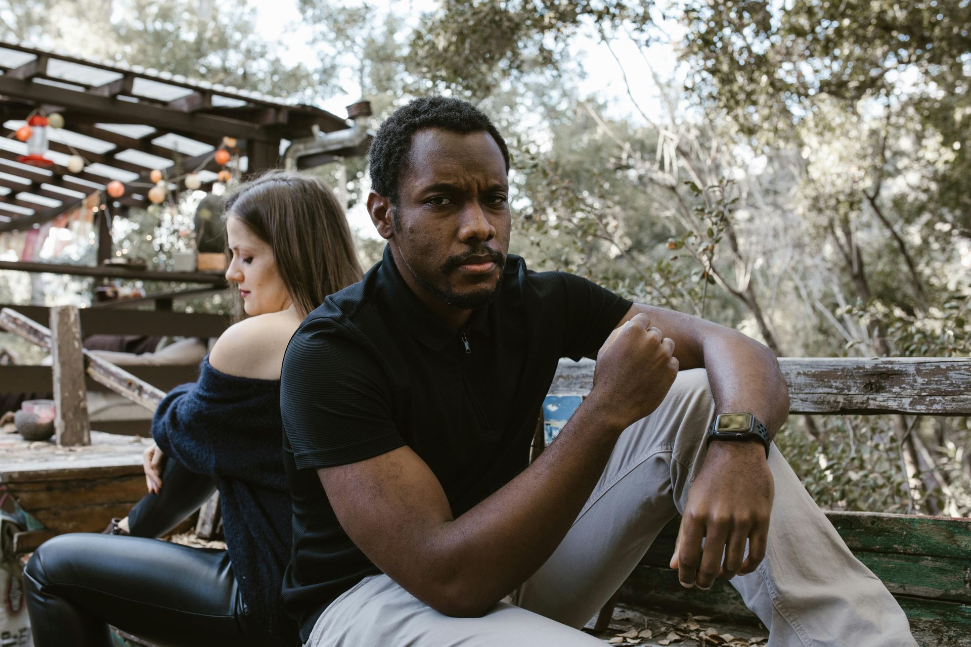 Man and woman sitting back to back outdoors, appearing upset. The man has a serious expression.