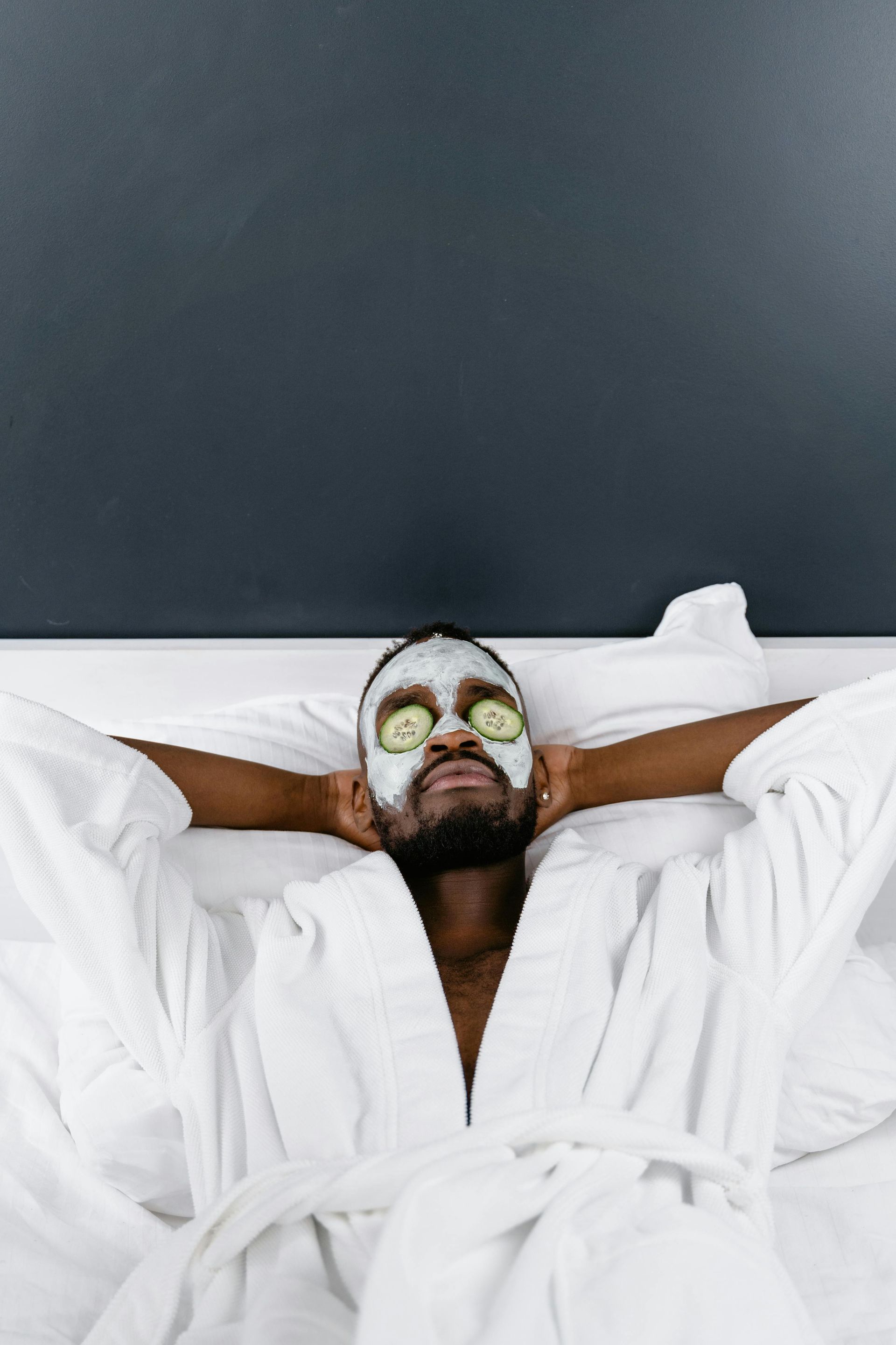 Man in white robe with face mask and cucumber slices on eyes, relaxing on bed.