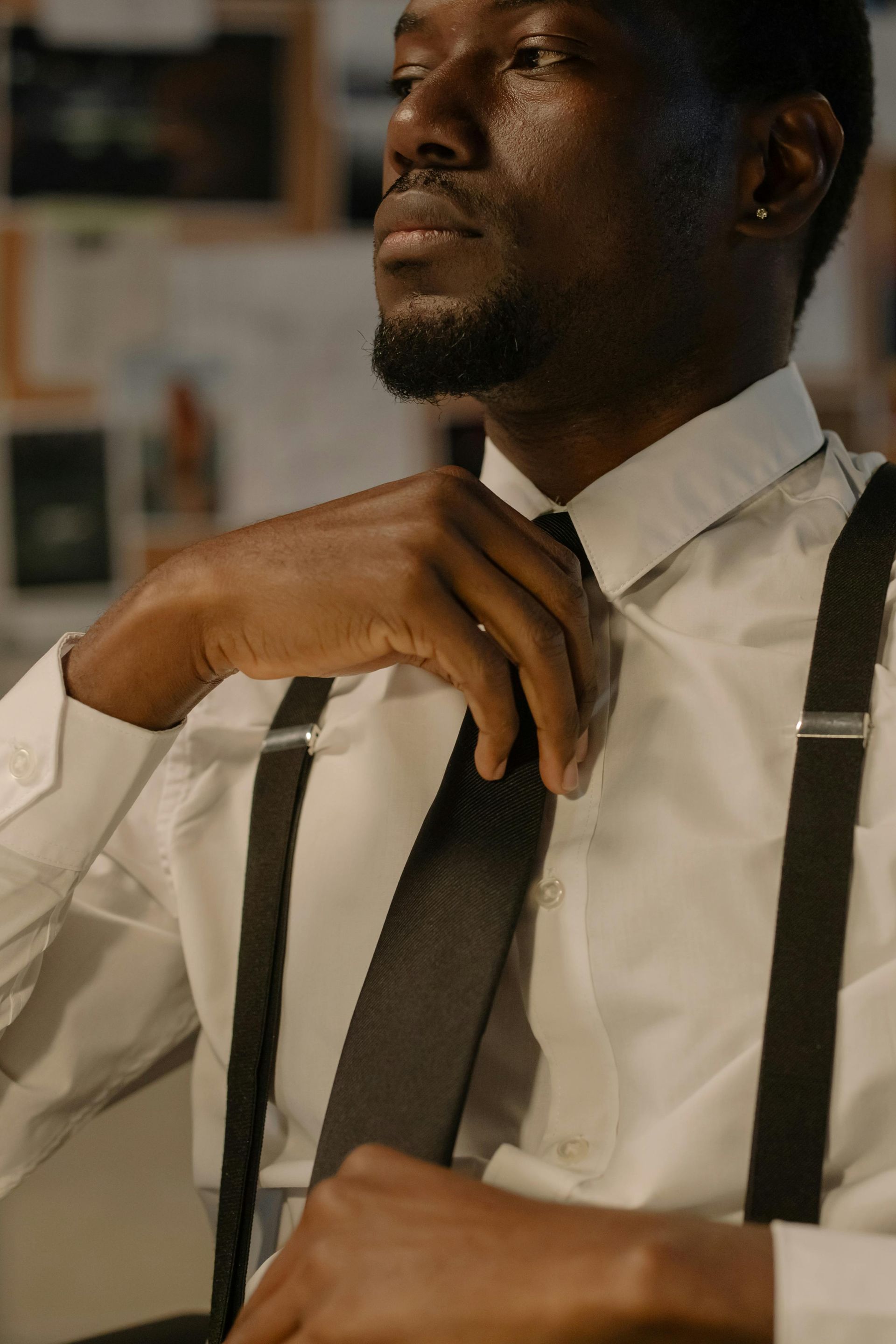 Man in white shirt, suspenders, adjusting tie; lit with soft light.