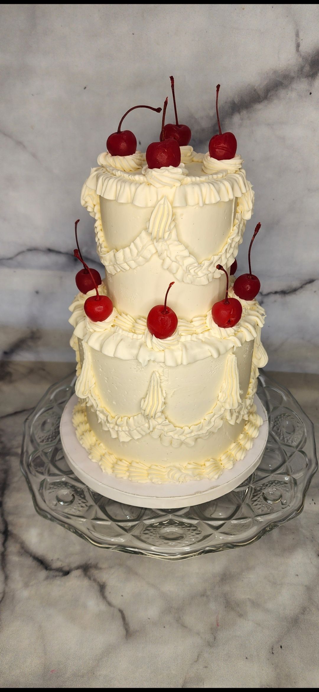 A two tiered cake with cherries on top is on a glass plate.