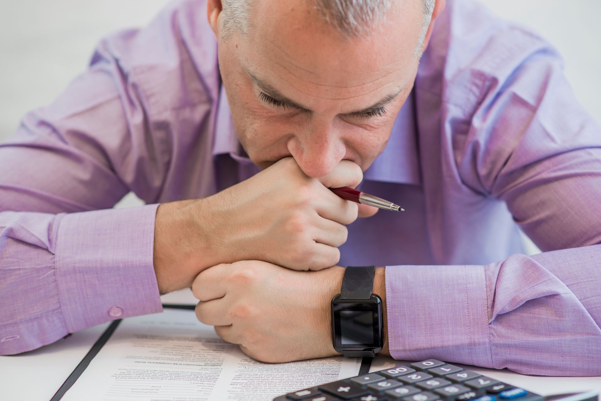 A man is sitting at a table with a calculator and a pen in his mouth.